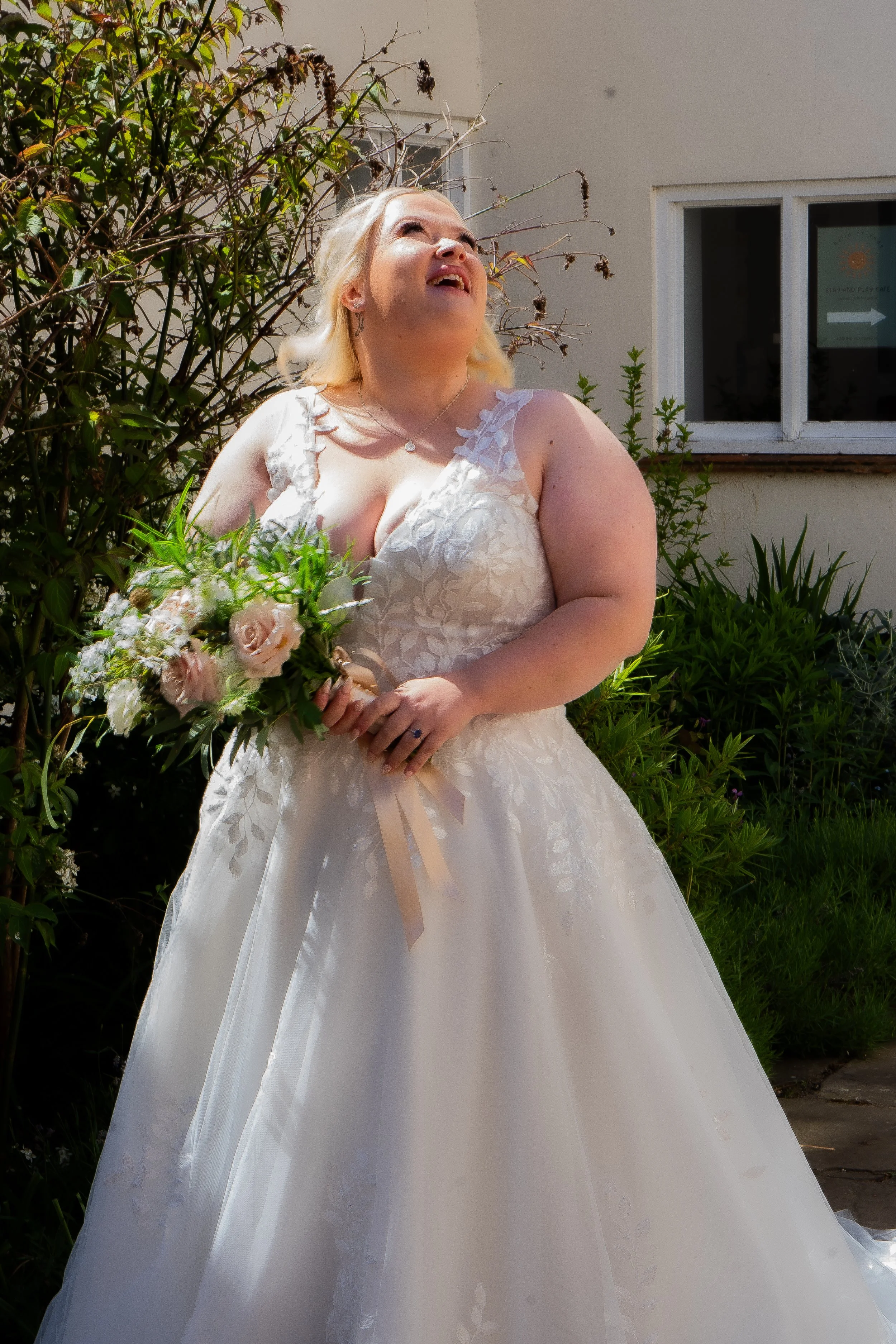 A woman in a white wedding dress holding a bouquet of flowers, smiling and looking upward outdoors near a building with a window and some plants.