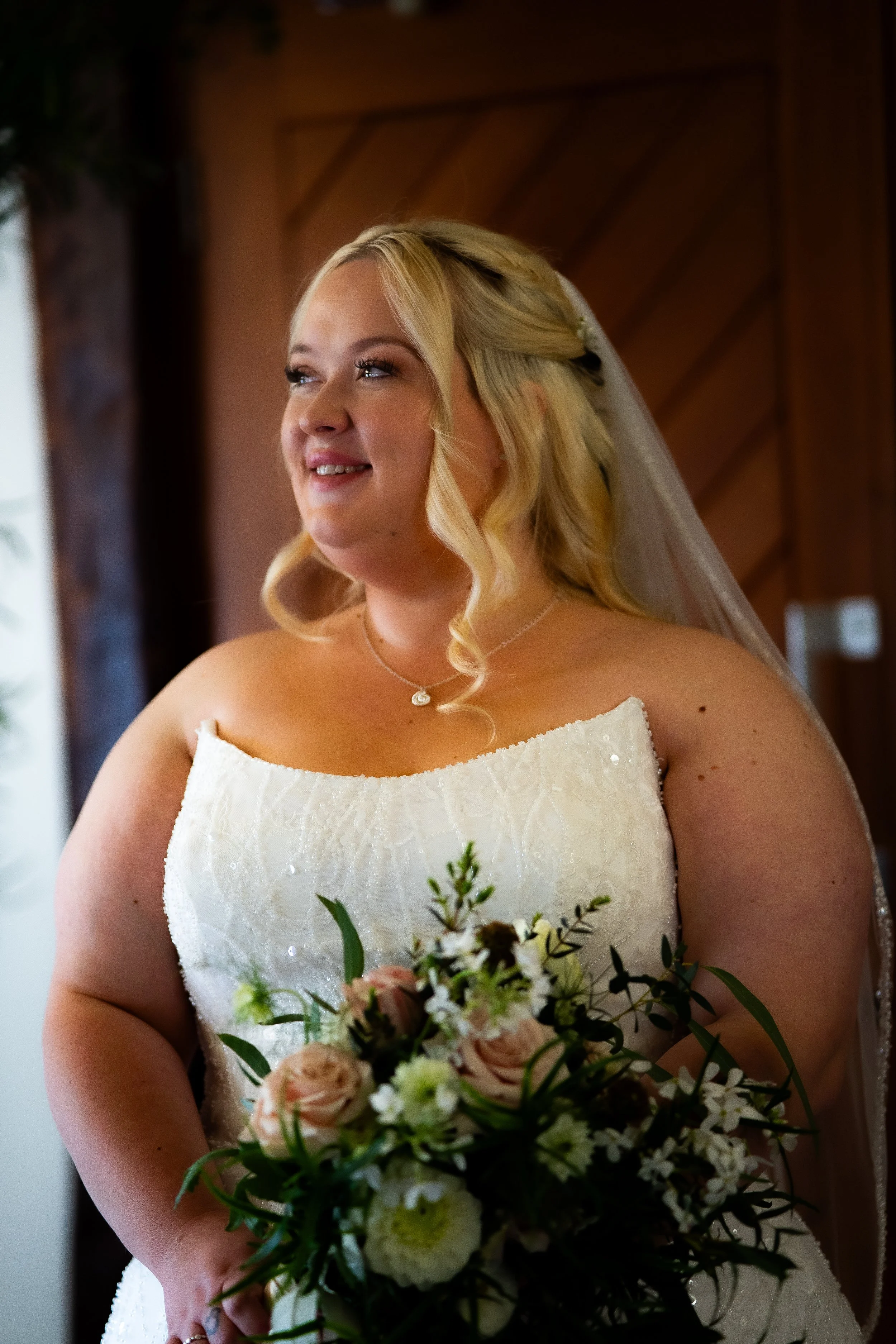 A bride wearing a white strapless wedding dress, holding a bouquet of flowers, smiling, with curly blonde hair and a veil in a wooden interior setting.
