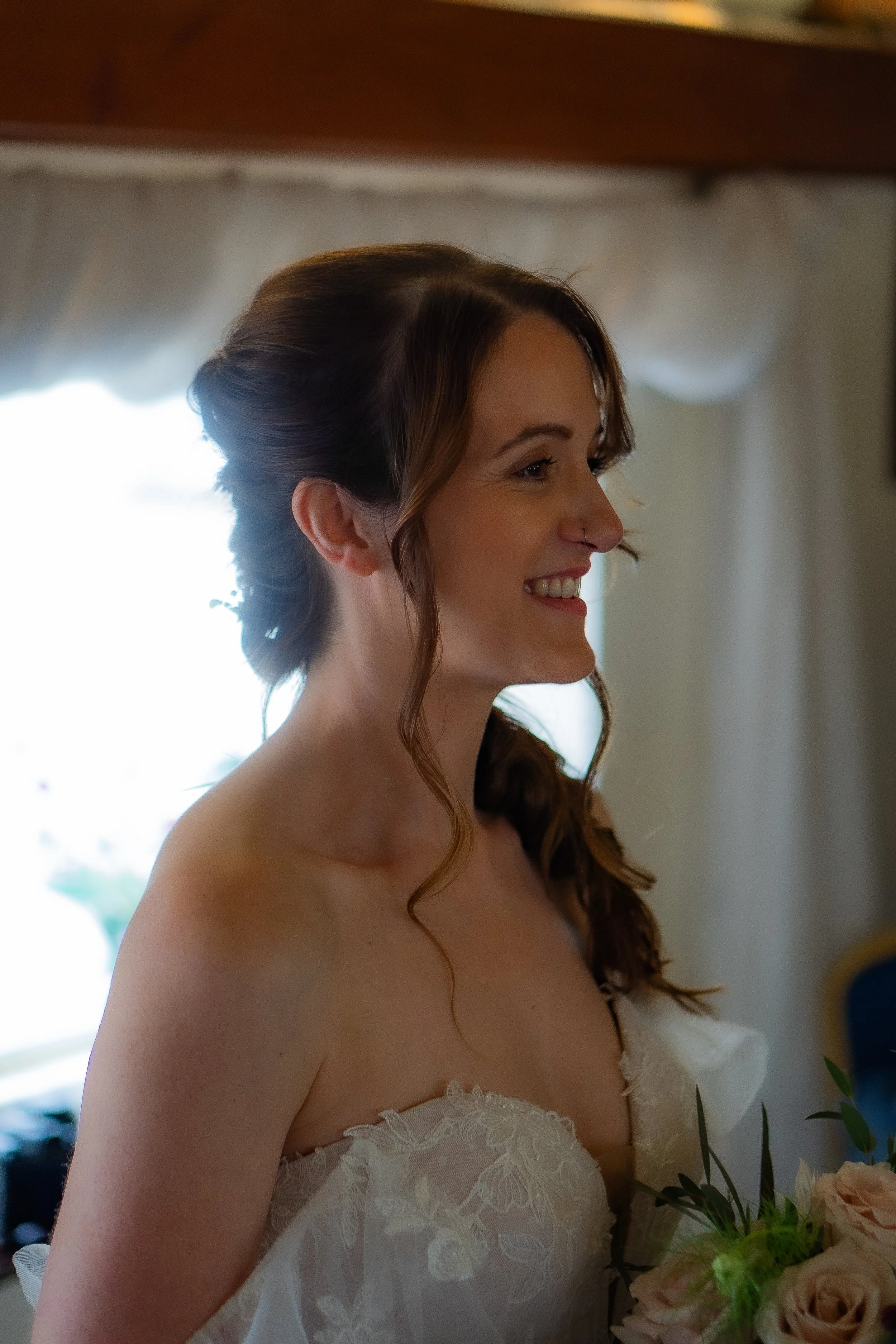 A smiling woman with brown hair styled in soft waves, wearing a strapless white lace dress, holding a bouquet of pale pink roses and greenery, standing indoors with a blurred background and natural light.