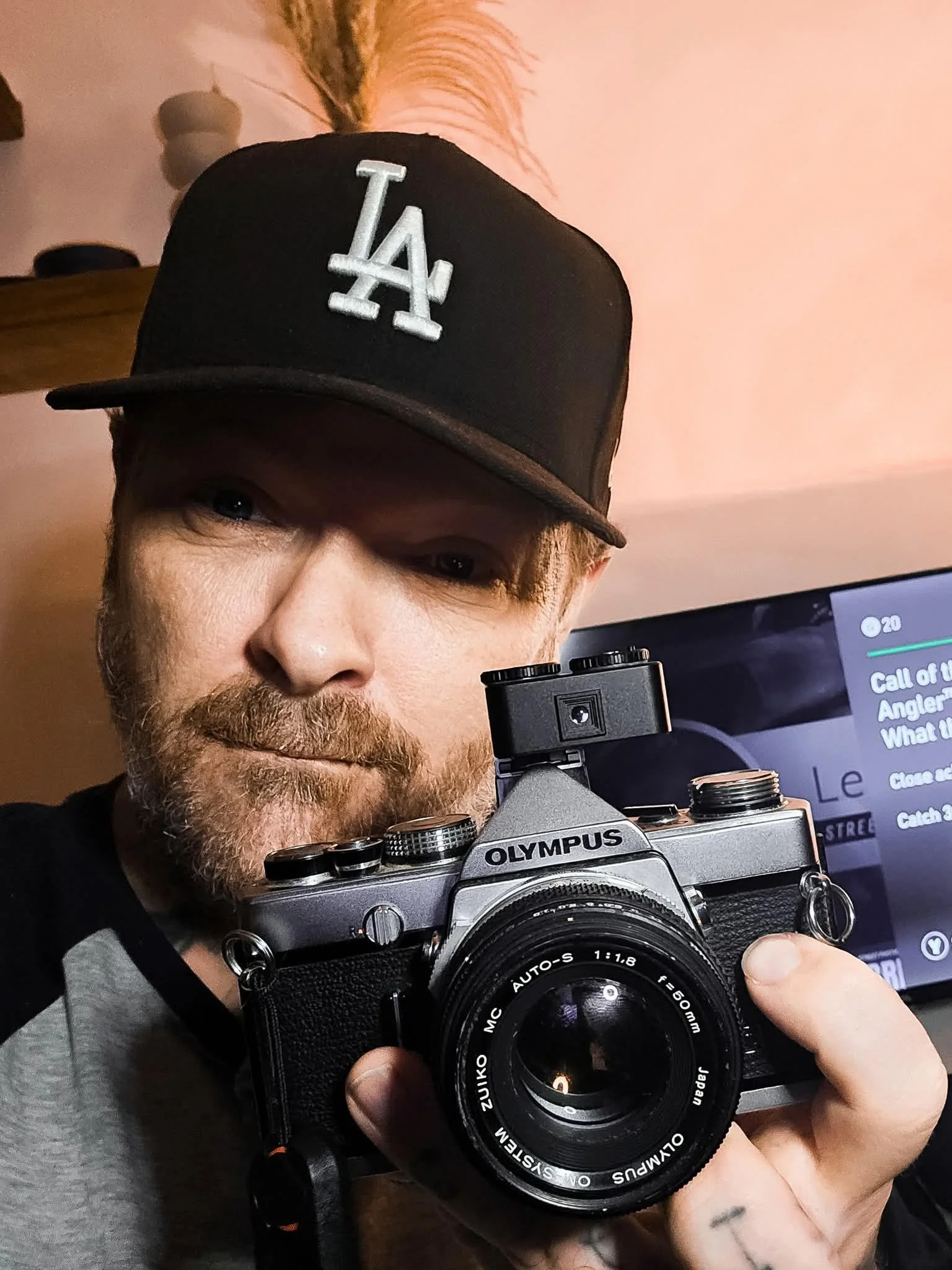 A man taking a selfie in a mirror, wearing a black cap with an LA logo, holding an Olympus camera with a zoom lens, with a computer screen in the background showing a music playlist or video.
