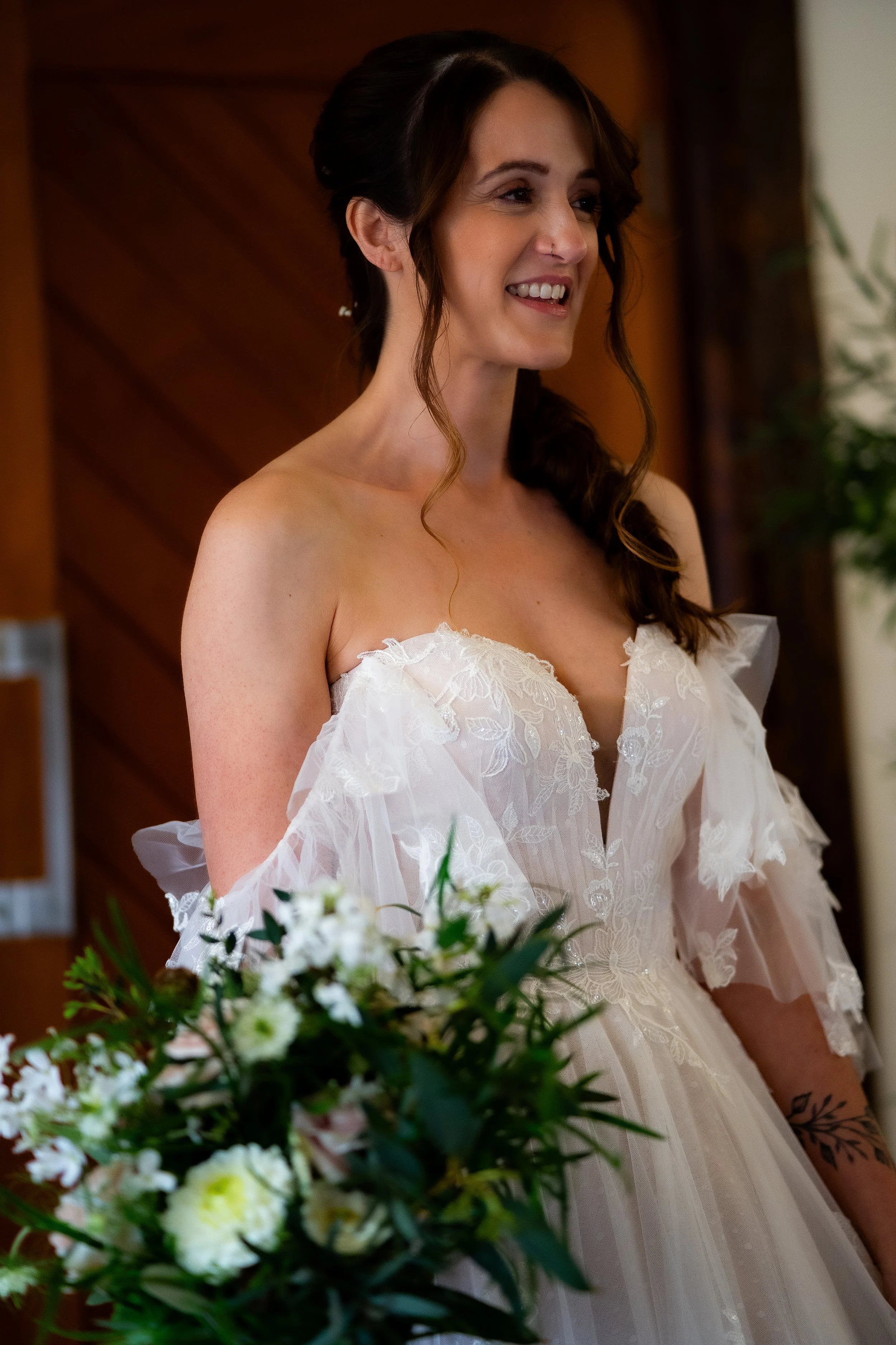 A smiling woman in a wedding dress standing indoors with a floral bouquet in front of her and a dark wood-paneled background.