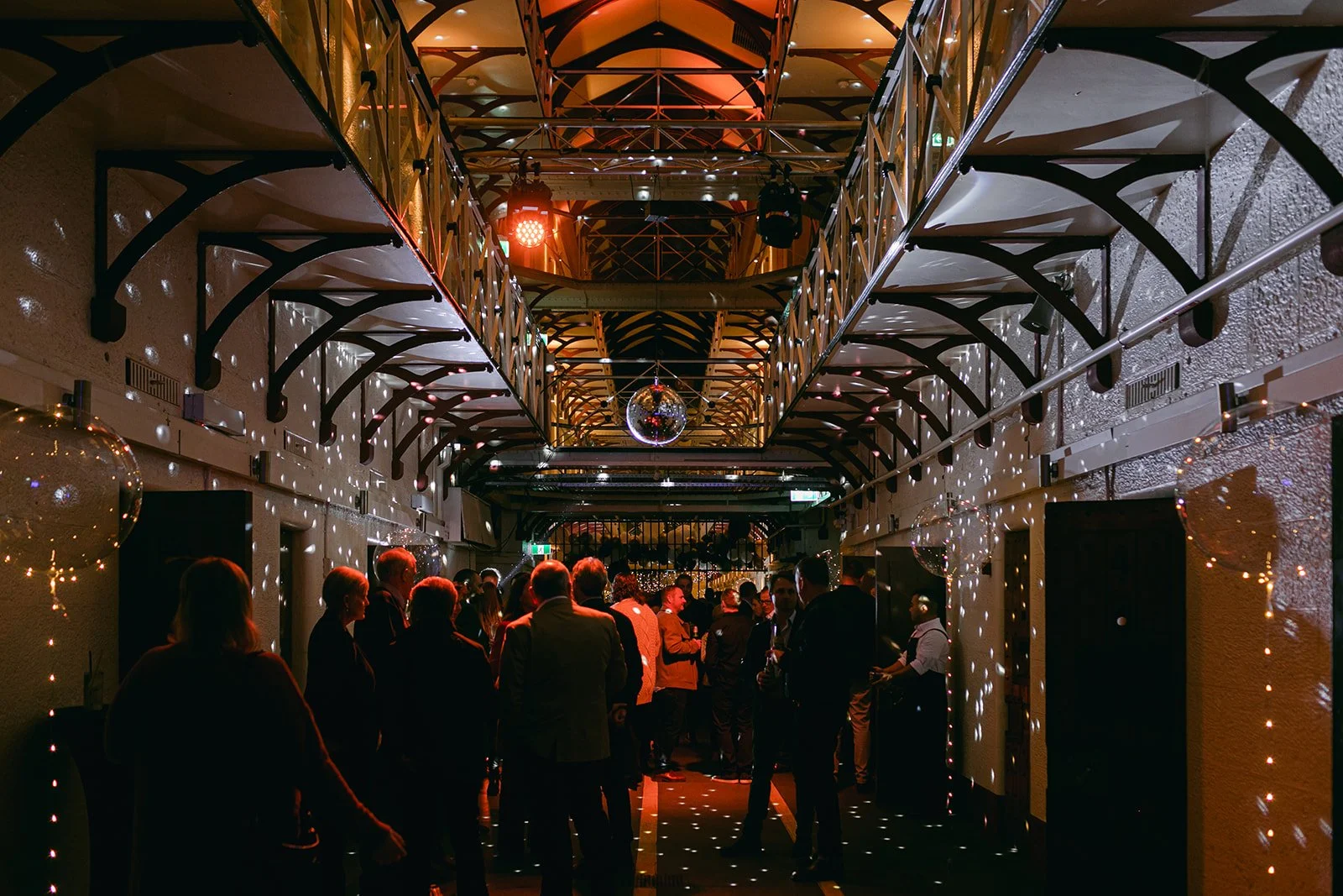 People socializing at an indoor party with festive lighting, a disco ball, and hanging decorations in a two-story venue.