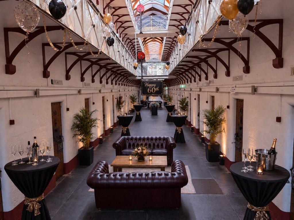 An indoor event space decorated with black, gold, and white balloons, with tables holding champagne bottles and glasses, and a sign that reads 'Happy 60th' in the background.