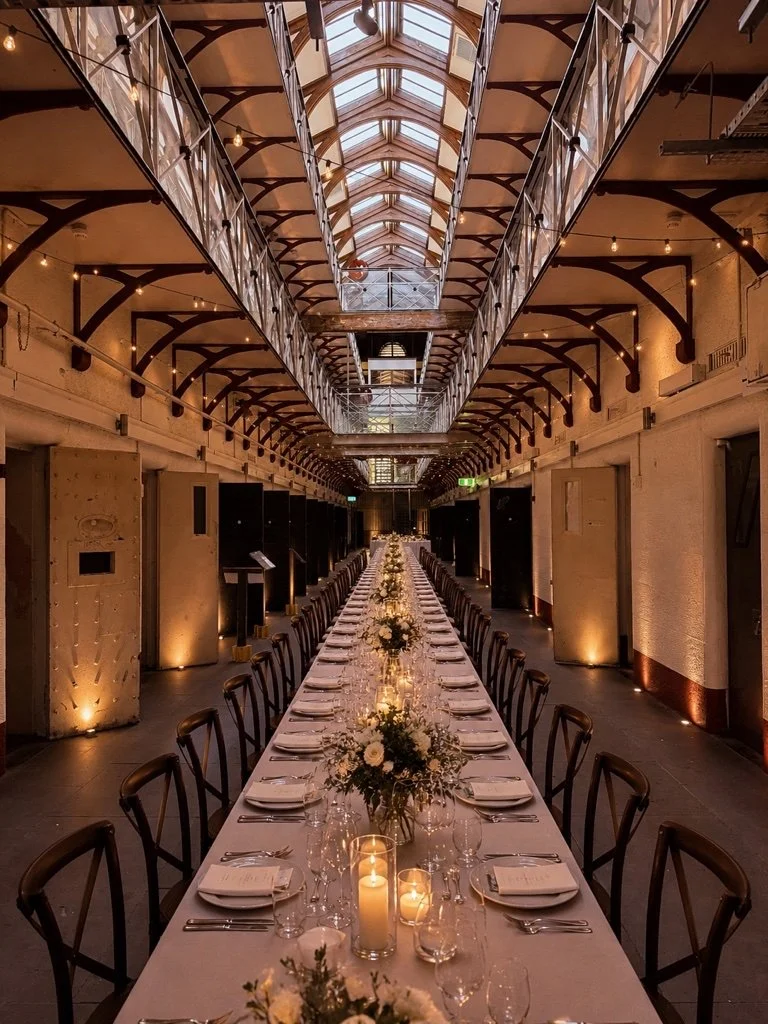 Long banquet table with white tablecloth, floral centerpieces, candles, and place settings in a warmly lit rustic-industrial event space with a high, arched glass ceiling.
