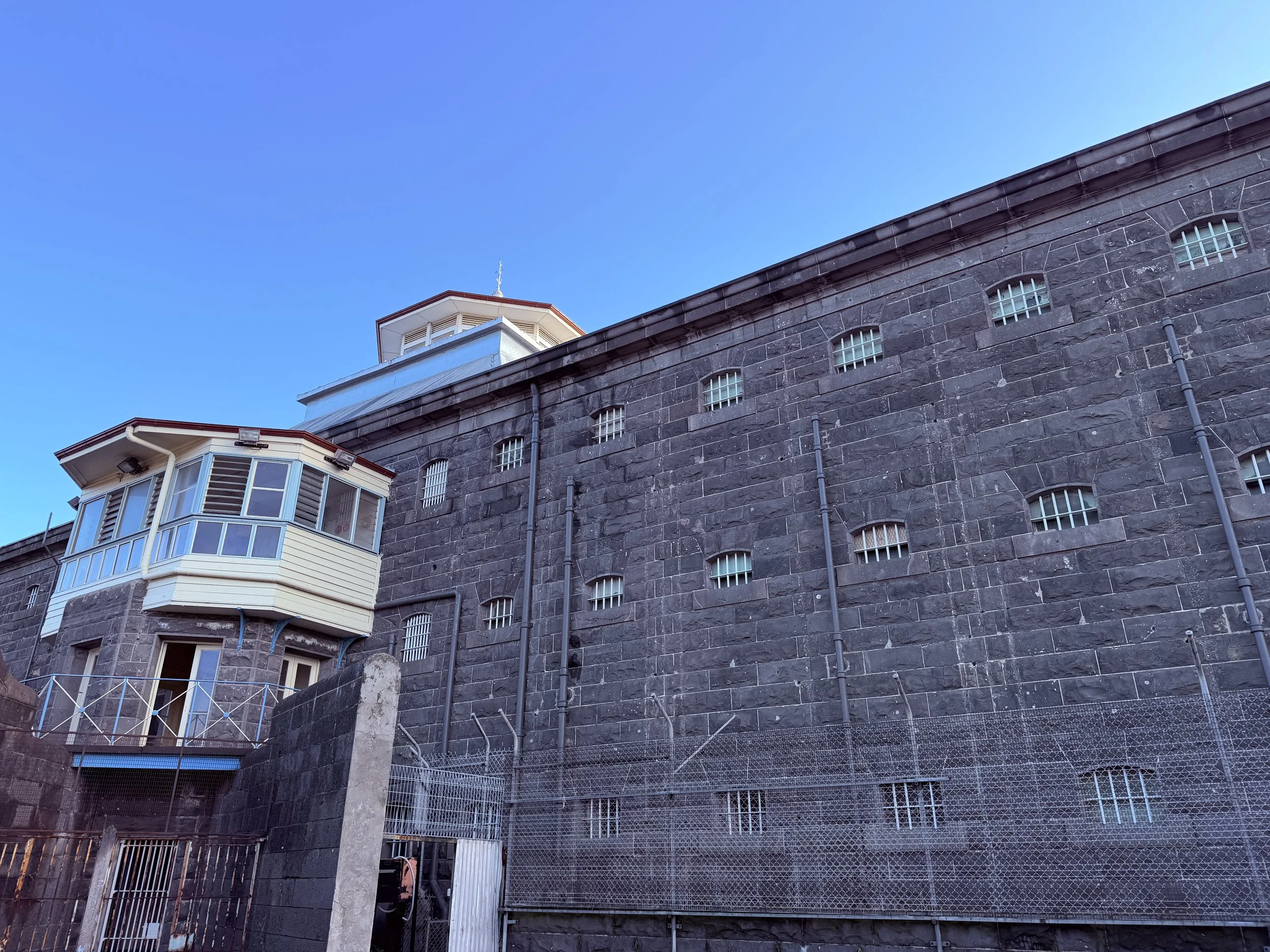 A stone building with barred windows, a balcony enclosed with wire fencing, and a tower at the top against a clear blue sky.