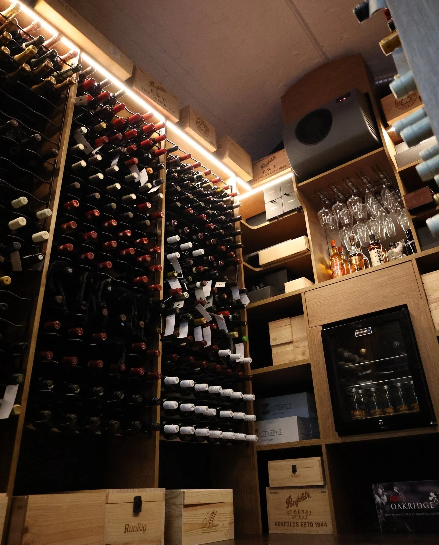 Wine bottles stored on wooden racks, with wine glasses and liquor bottles on shelves, and a small wine cooler in a cozy cellar or wine room.