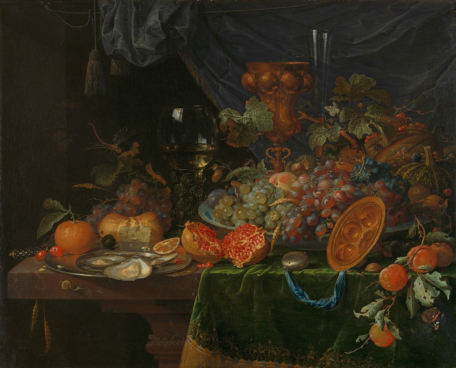 A Baroque-style still life painting featuring a variety of fruits, grapes, pomegranate, apple, orange, and pomegranate seeds. There are also shells, a glass goblet, a small ornate box, a glass vase, and decorative objects including a yellow glass plate and a carved wooden statue. The table is draped with green fabric and a dark cloth background with a blue drape and tassel.