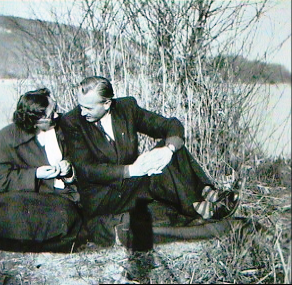 Two women in vintage clothing sitting on a bench by a body of water, engaged in conversation.