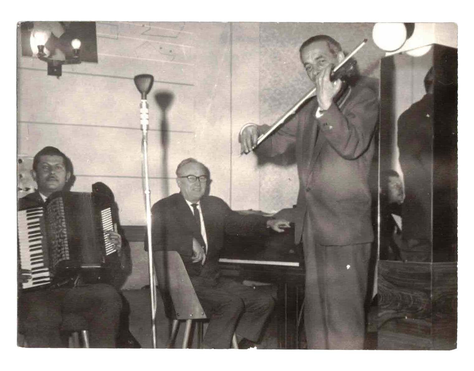 A black and white photo of a man playing the flute, a man playing the accordion, and a man sitting at a piano in a room with a wood-paneled wall and a hanging light fixture.