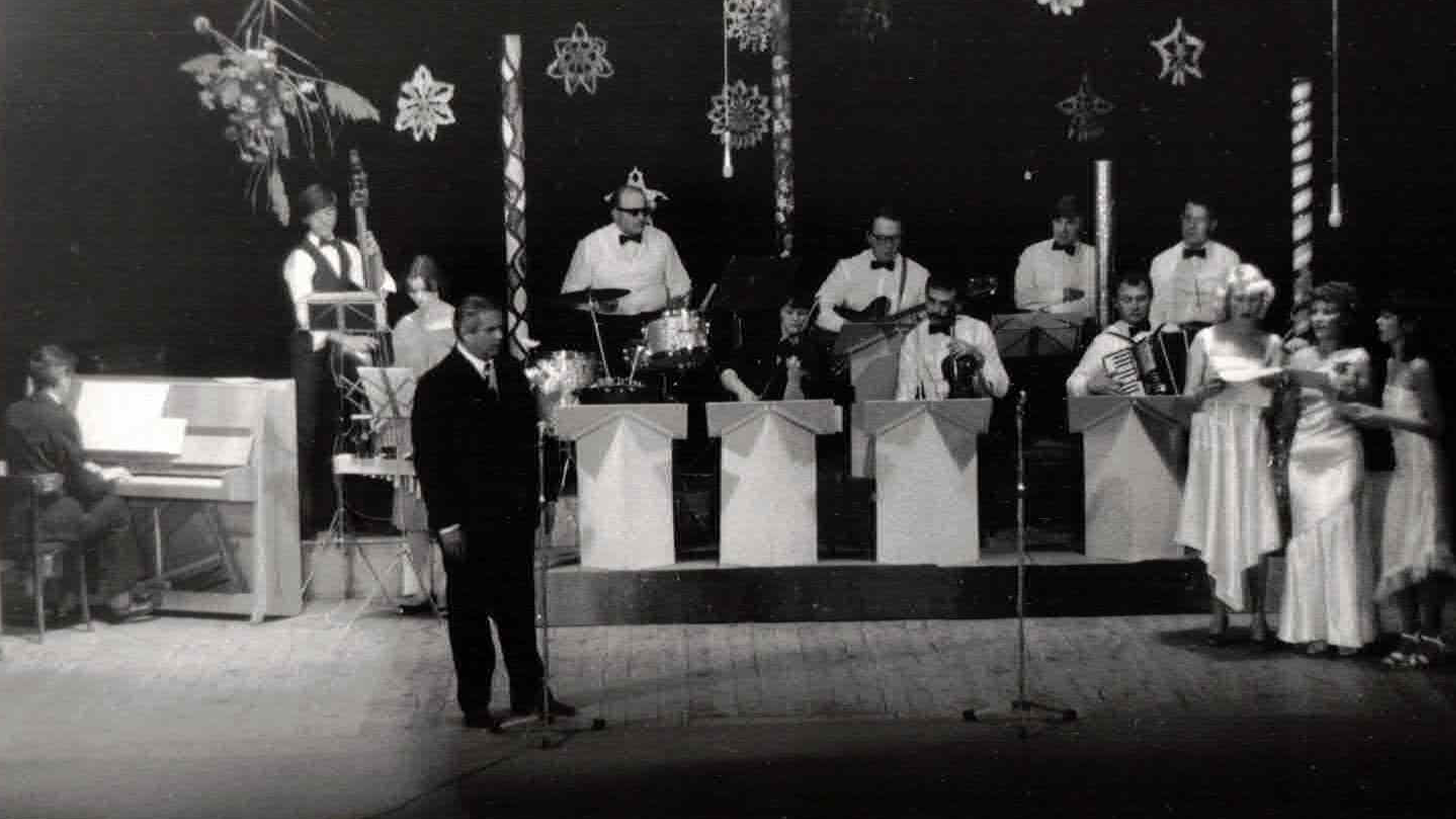 A vintage black and white photo of a band performing on stage with Christmas decorations, including snowflakes and candy cane patterns, in the background. The band features a pianist, a singer, a drummer, a guitarist, a saxophonist, an accordion player, and a group of women singing.