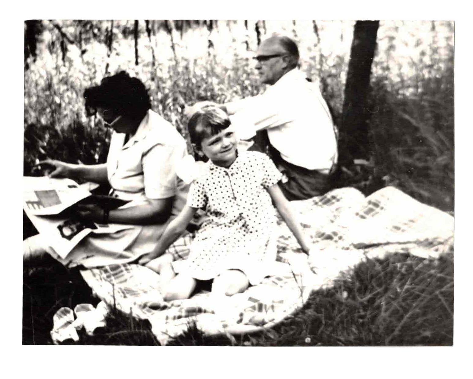 Black and white photo of three people sitting on a blanket outdoors. One woman is reading a book, a young girl with short hair and a polka dot dress is sitting on the blanket, and an older man with glasses is sitting behind her. They are surrounded by trees.