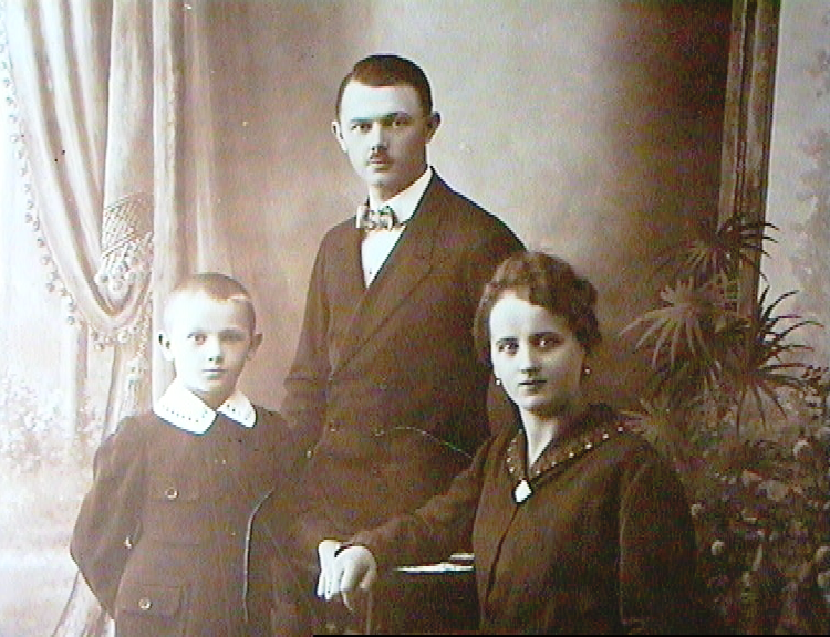 A black-and-white portrait of a family with a man, woman, and a young girl, dressed in early 20th-century clothing, posed indoors with curtains and a potted plant in the background.
