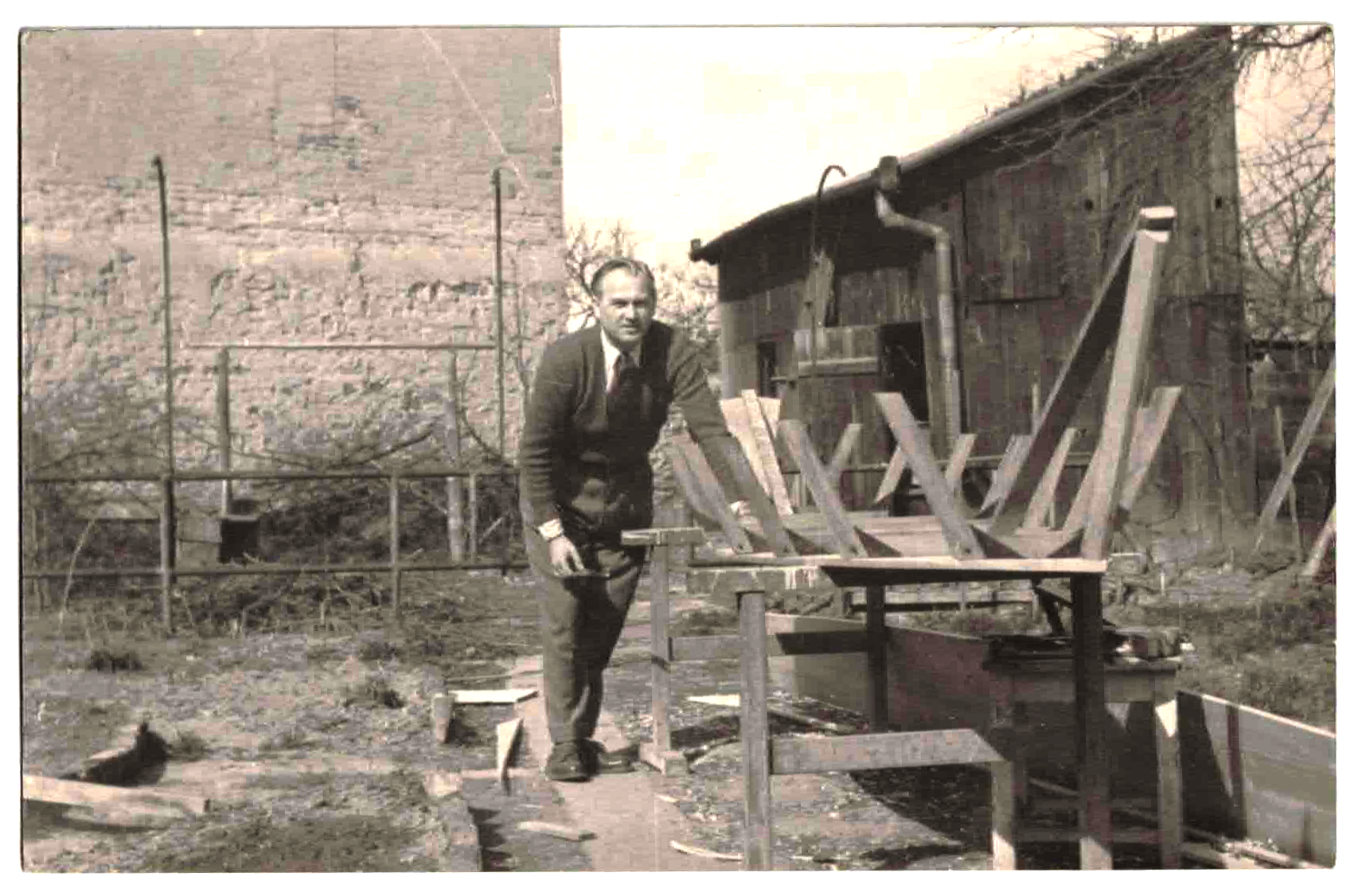 Black and white photo of a man working on a woodworking project outdoors in a backyard, with a small wooden house or shed in the background and some trees and fencing around.