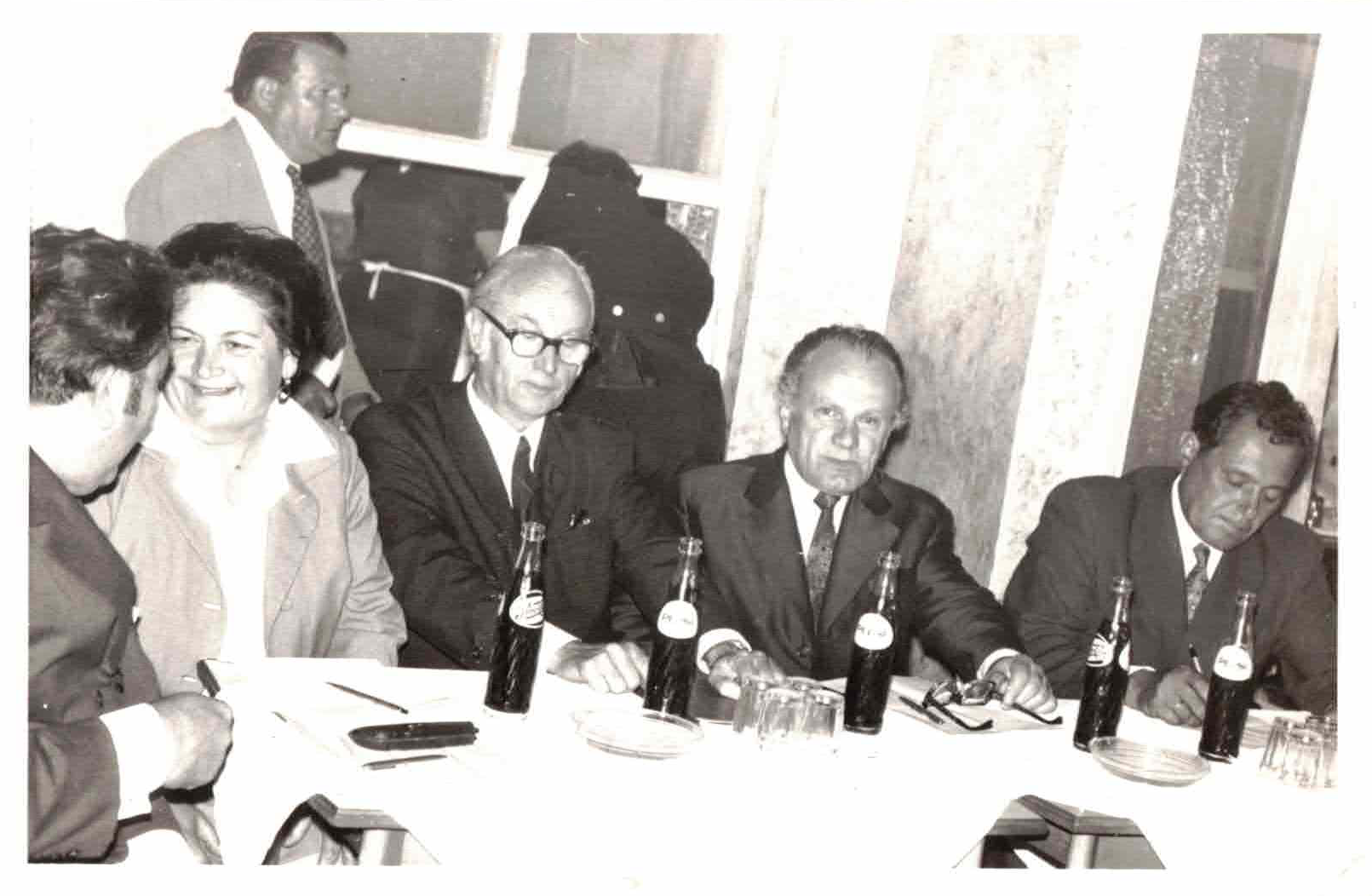 Black and white photo of a group of people sitting at a table during a meeting or social event. Four men are seated, with bottles of beer in front of them, and a woman is standing and talking to two women seated at the table. The setting appears to be indoors.