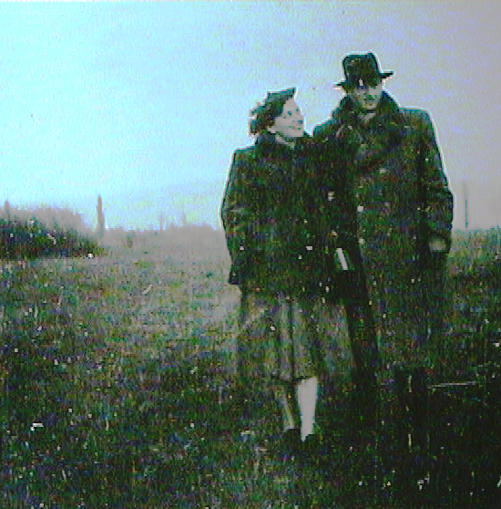 Black and white photo of a man and woman in historical clothing standing outdoors, the woman looking at the man while he looks into the distance.