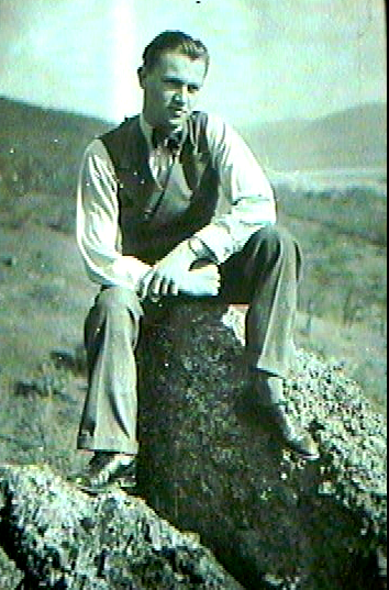 A man with slicked-back hair wearing a vest over a collared shirt, sitting on a rock or large stump in an open grassy field with hills in the background.