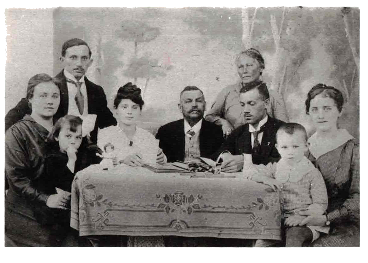 A black-and-white family portrait showing nine people gathered around a table with a embroidered tablecloth. Four women, three men, and two children are present, with some seated and others standing behind. The scene has a plain backdrop with painted scenery.