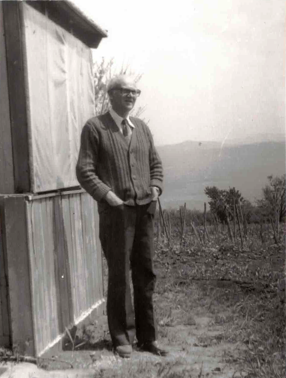 A man with light-colored, spiky hair and glasses stands outdoors beside a rustic shed, with a landscape of hills and sparse vegetation in the background.