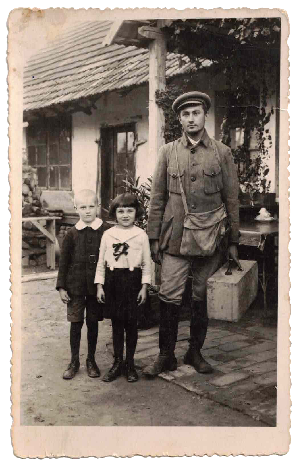 A black-and-white photograph of a soldier standing outdoors with two children, one boy and one girl, in front of a house with a tile roof, window, and door. The scene appears to be from an earlier time period, possibly wartime.