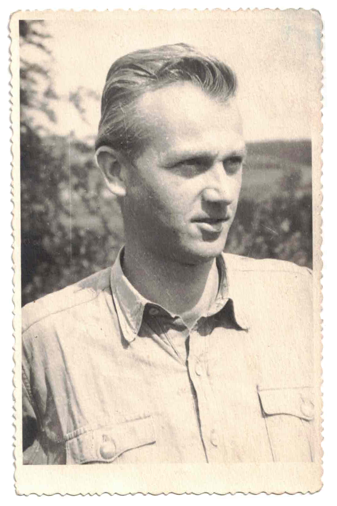 Black and white portrait of a young man with neatly combed hair, wearing a button-up shirt with pockets, outdoors with trees and sky in the background.
