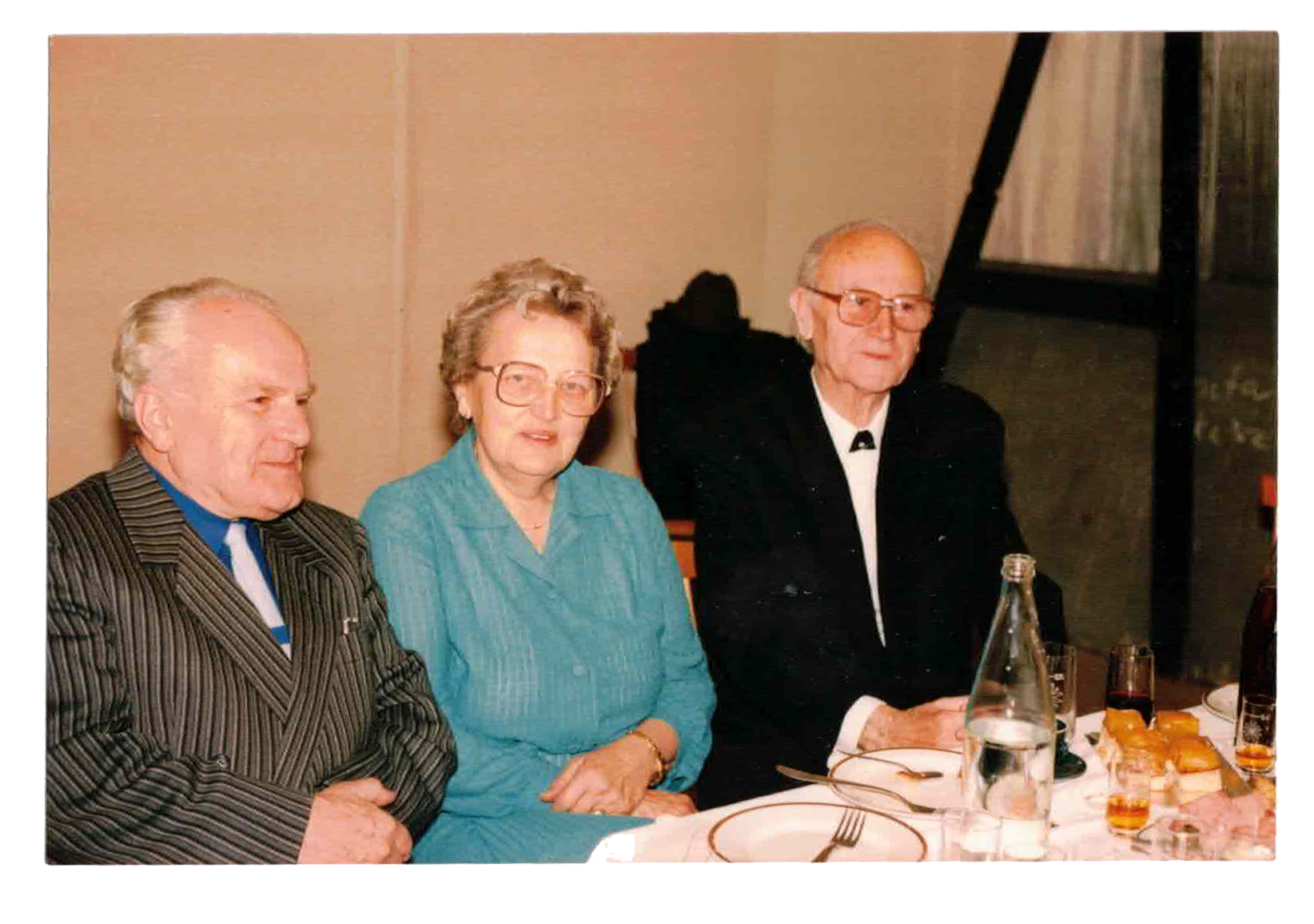 Three elderly people sitting at a dinner table with drinks and food, dressed in formal attire, indoors with beige walls and a black metal staircase in the background.