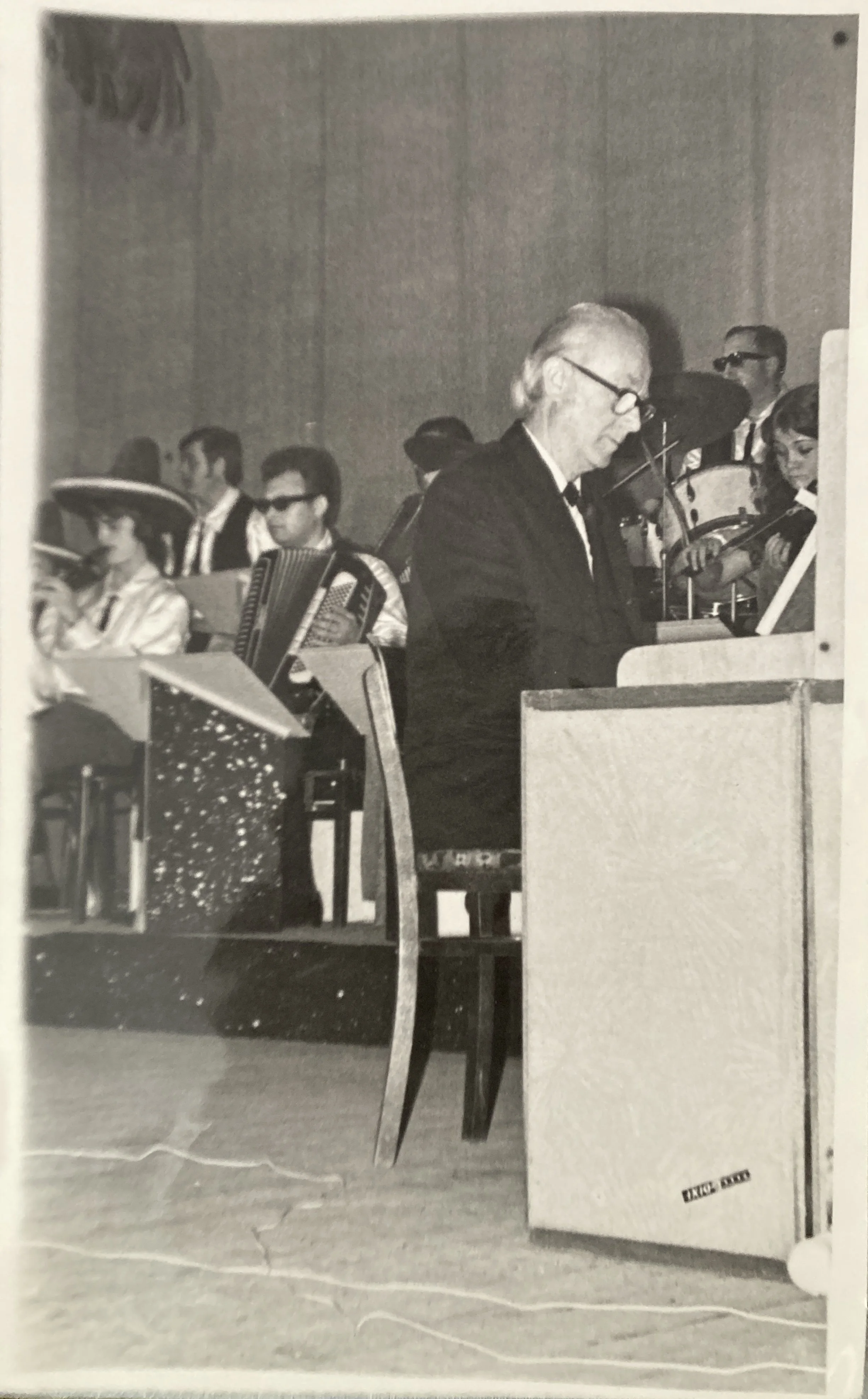 Black and white photograph of a man with glasses playing a piano in a room with several seated and standing people, possibly at a social event or gathering.