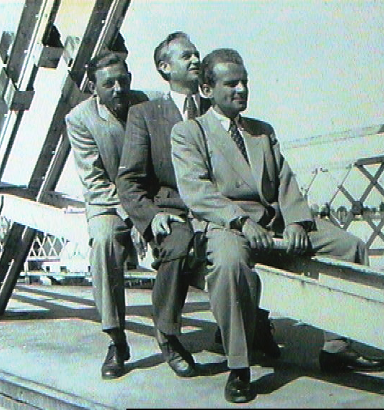 Three men in suits sitting on a carnival ride or amusement park ride, smiling and looking to the right.