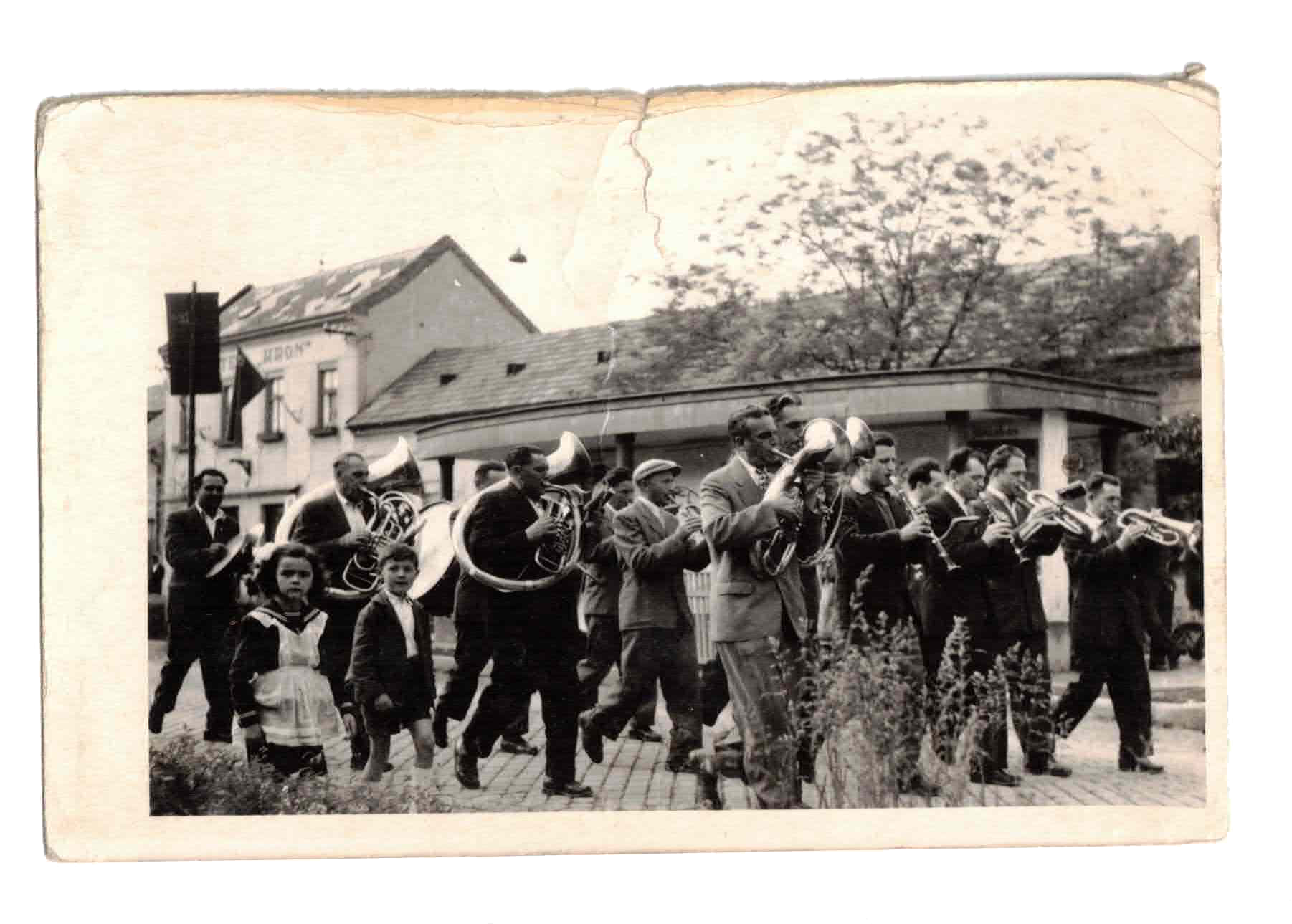 A black and white photo of a marching band playing musical instruments outdoors, including children and adults, with a building, trees, and a lamppost in the background.