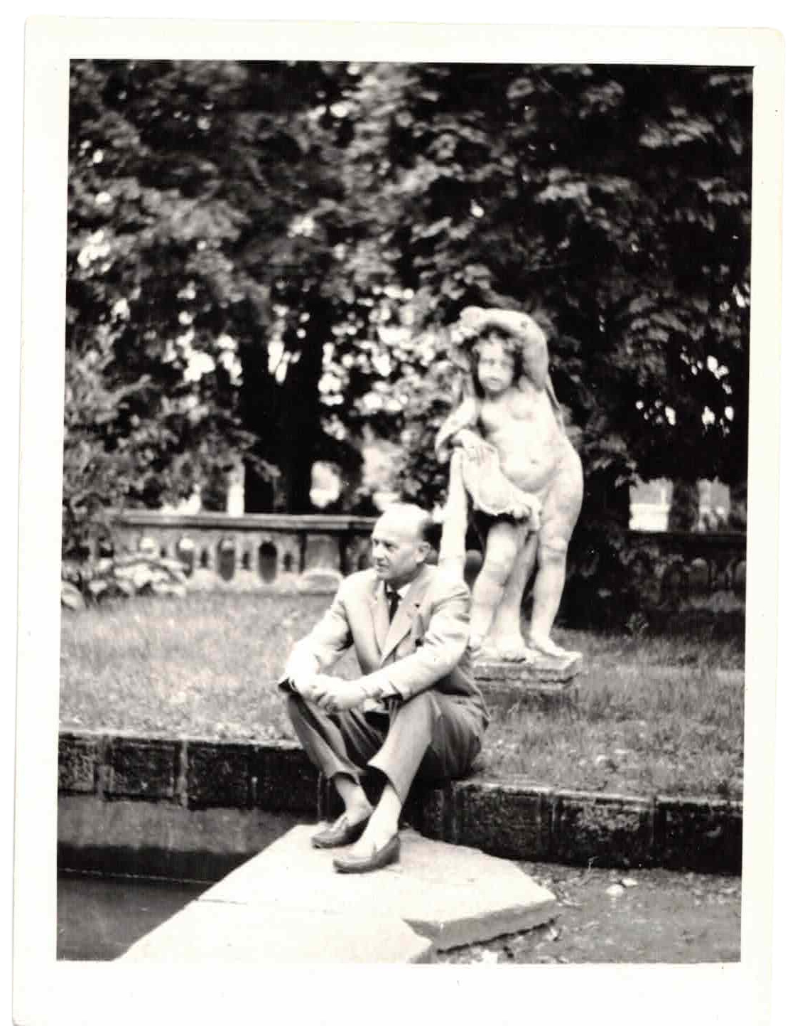 A black and white photograph of a man sitting on a stone ledge with a sculpture of a woman behind him, set in a garden with trees and a stone fence.