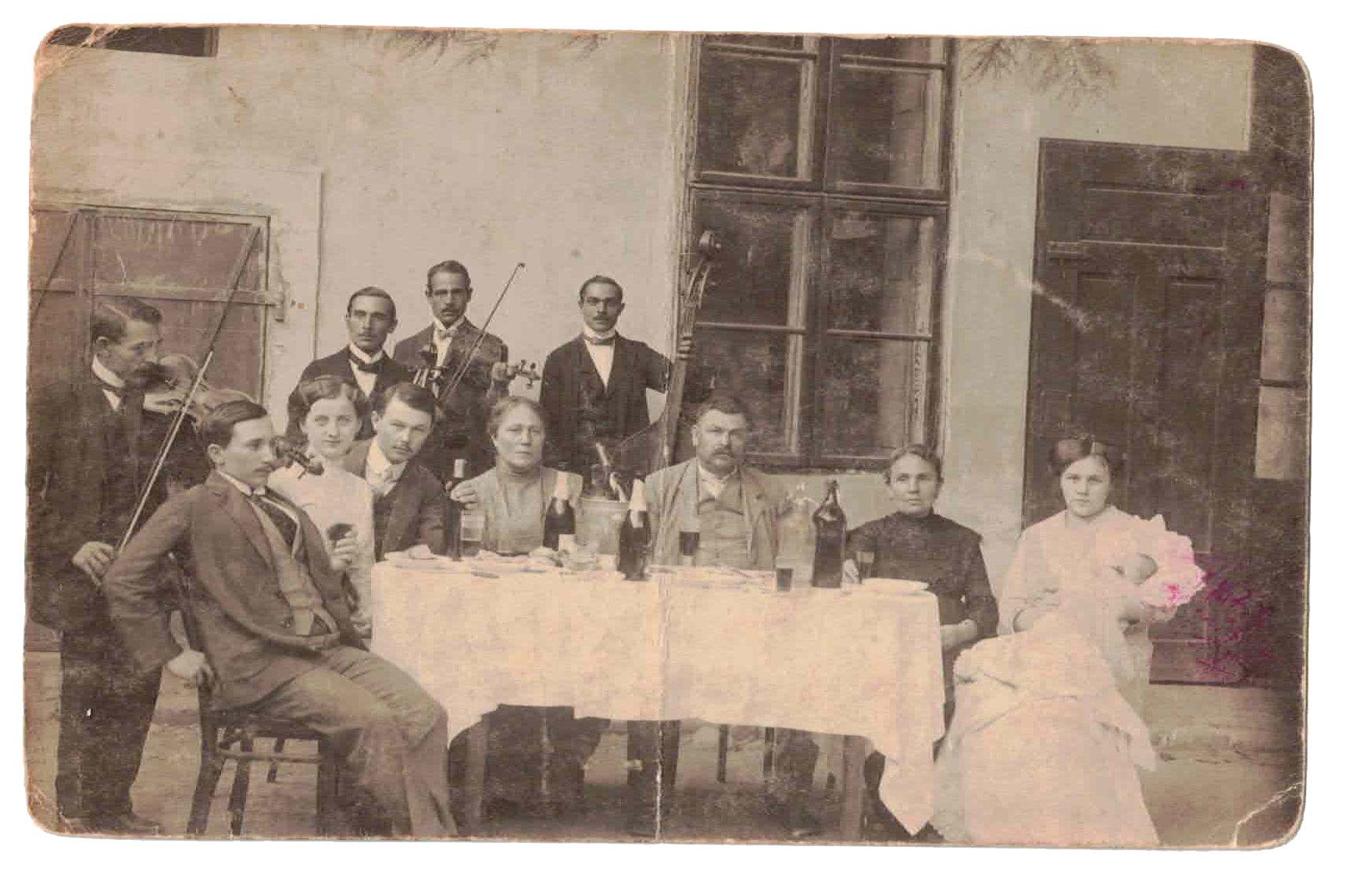 A vintage photograph of eleven people, six men, three women, and two children, gathered around a table outdoors in front of a building with an open window. Some men are holding musical instruments, such as violins, and there are bottles and glasses on the table, indicating a social gathering or celebration.