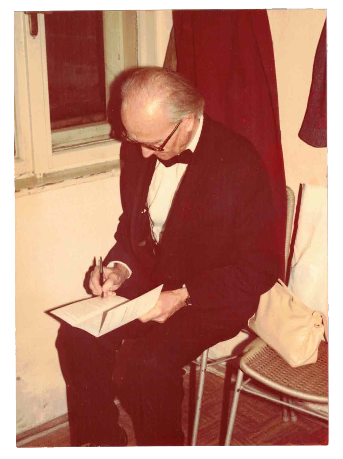 An elderly man dressed in a tuxedo with a bow tie, sitting on a chair, writing on a piece of paper or a card, with glasses and receding hair, in a room with a window and a beige bag on a chair.