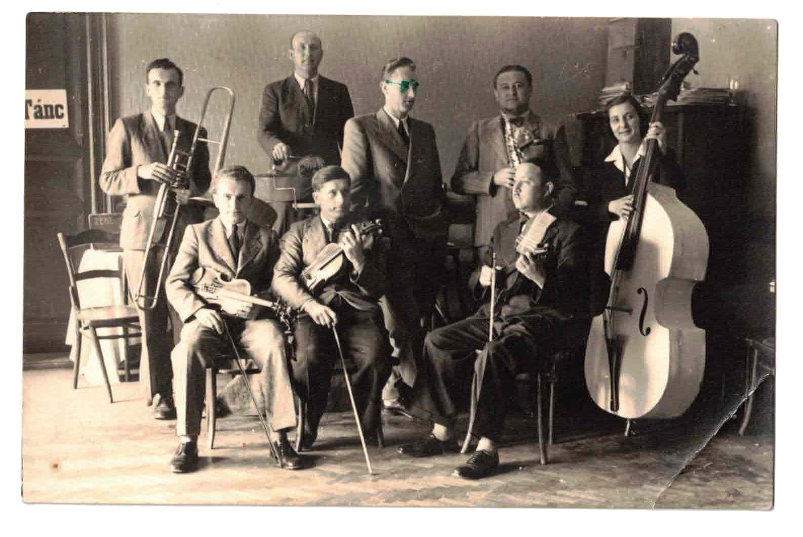 Black and white photo of a band with nine members holding musical instruments, including a double bass, violin, trombone, and clarinet, in a room with a plain wall background.