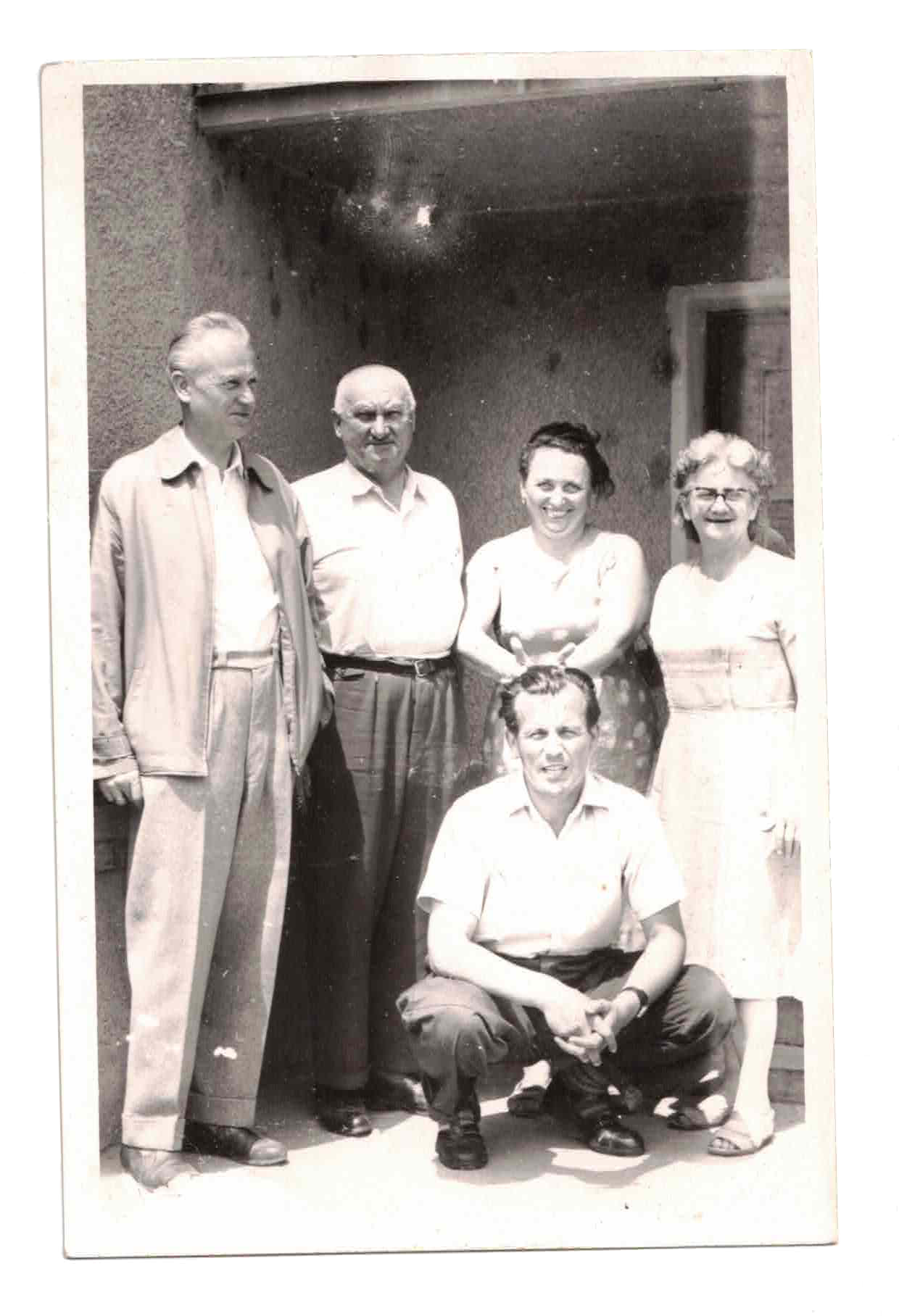 Black and white photo of six people, five standing and one sitting in front. They are outside near a wall, smiling and posing for the picture.