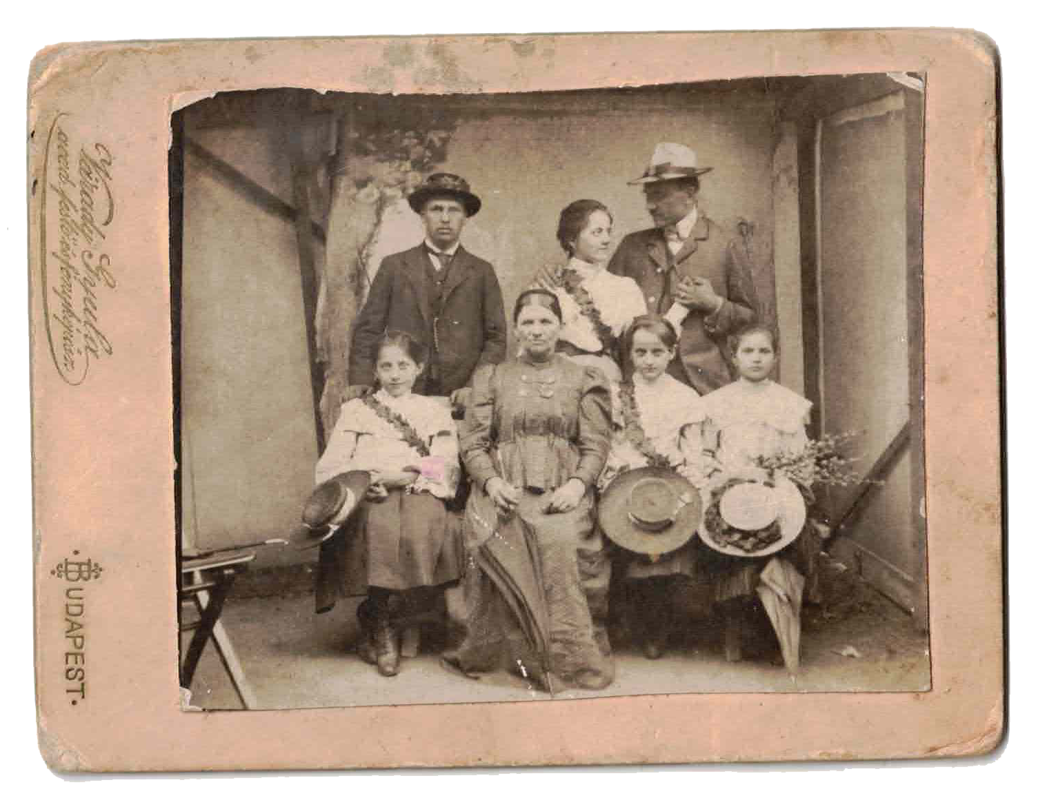A vintage black-and-white group photo featuring a man, a woman, four girls, and a boy, posed outdoors with trees in the background. The man and woman are at the center, with the children around them, some holding hats and flowers.