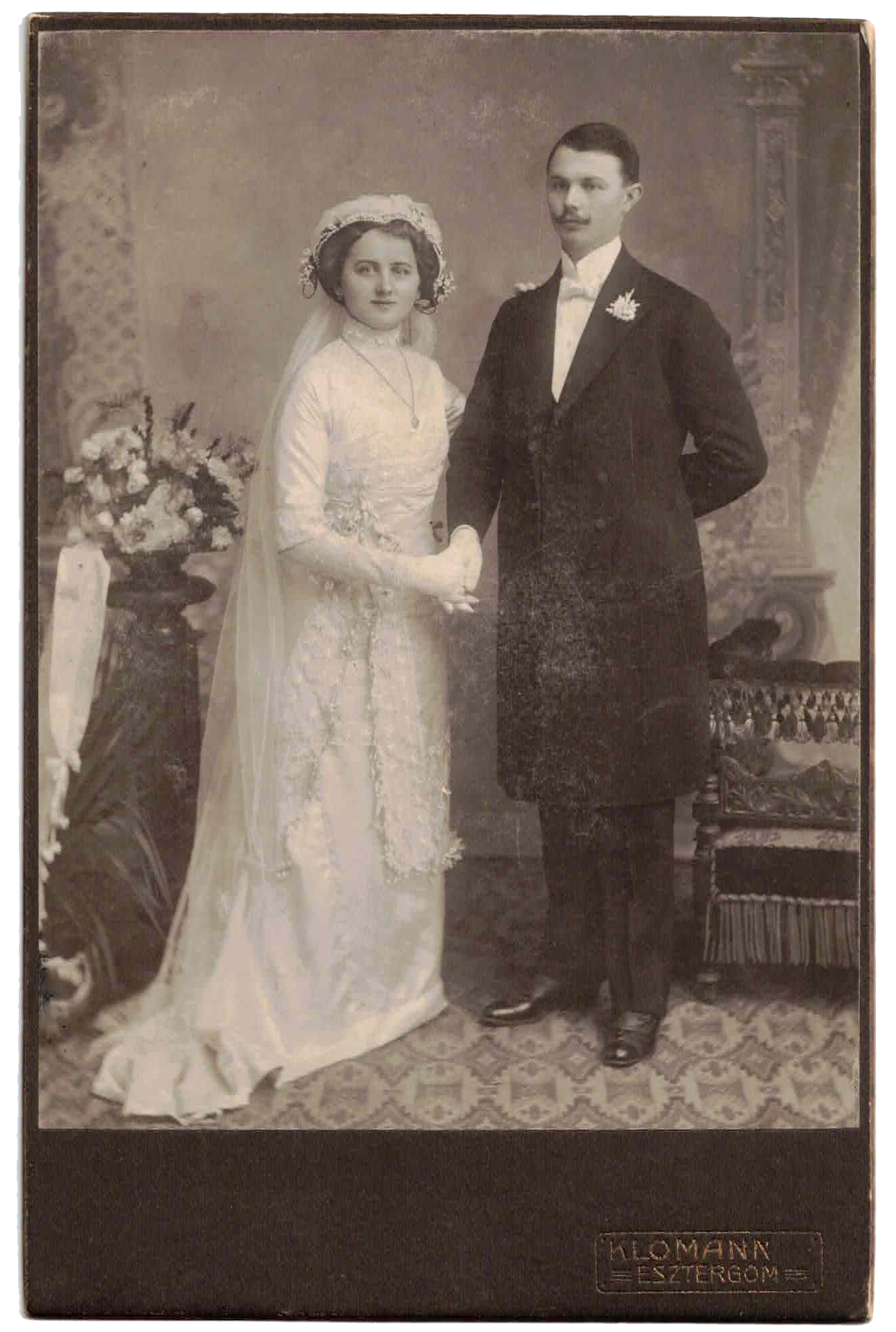 A vintage black and white photograph of a bride and groom standing together indoors. The bride is wearing a long white wedding gown with lace details, gloves, a veil, and a headpiece. The groom is dressed in a dark suit with a white shirt, bow tie, and a floral boutonniere. They are holding hands, and there are decorative vases with flowers and an ornate backdrop in the background.