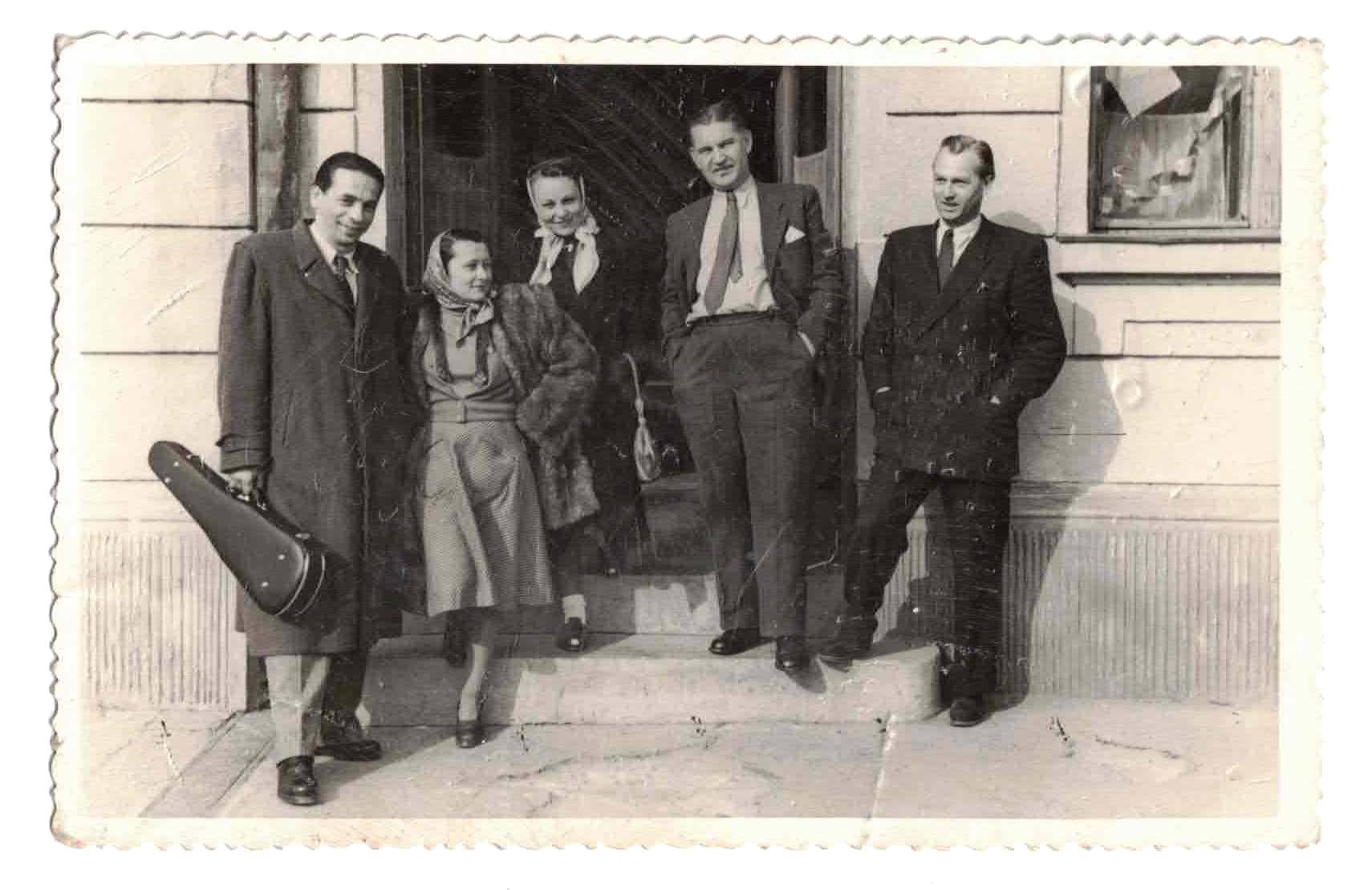 Old black and white photo of a family standing outside a building, with five people, including two men, one woman, one girl, and one boy, dressed in vintage clothing from the mid-20th century. The woman and girl are wearing headscarves, and the boy is holding a guitar case.