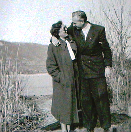 A black-and-white photo of a man and woman standing outdoors near a body of water, with trees and hills in the background. They are smiling and looking at each other, with the man embracing the woman.