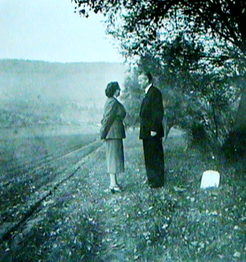A black-and-white photo of a man and woman standing on a grassy roadside near trees and bushes, facing each other.