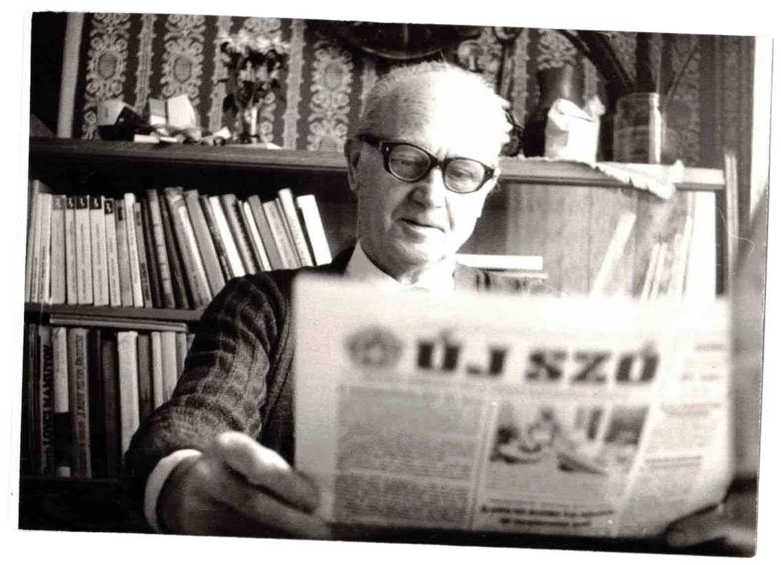 An elderly man with glasses reading a newspaper in a room with bookshelves and patterned wallpaper.