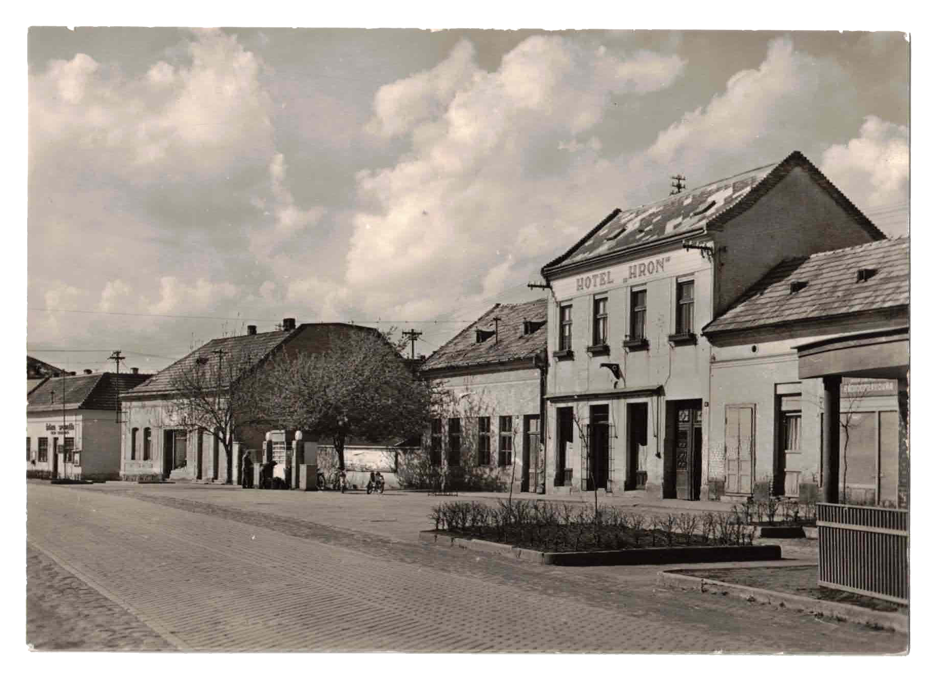 A historic small-town street scene in black and white, featuring a row of old buildings, including the Hotel Iron. The street appears quiet with only a few bicycles parked, and trees are lining the sidewalk under a partly cloudy sky.