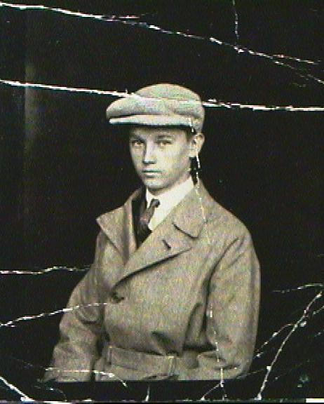 Black and white photograph of a young man wearing a flat cap and a coat, with a serious expression, against a dark background.