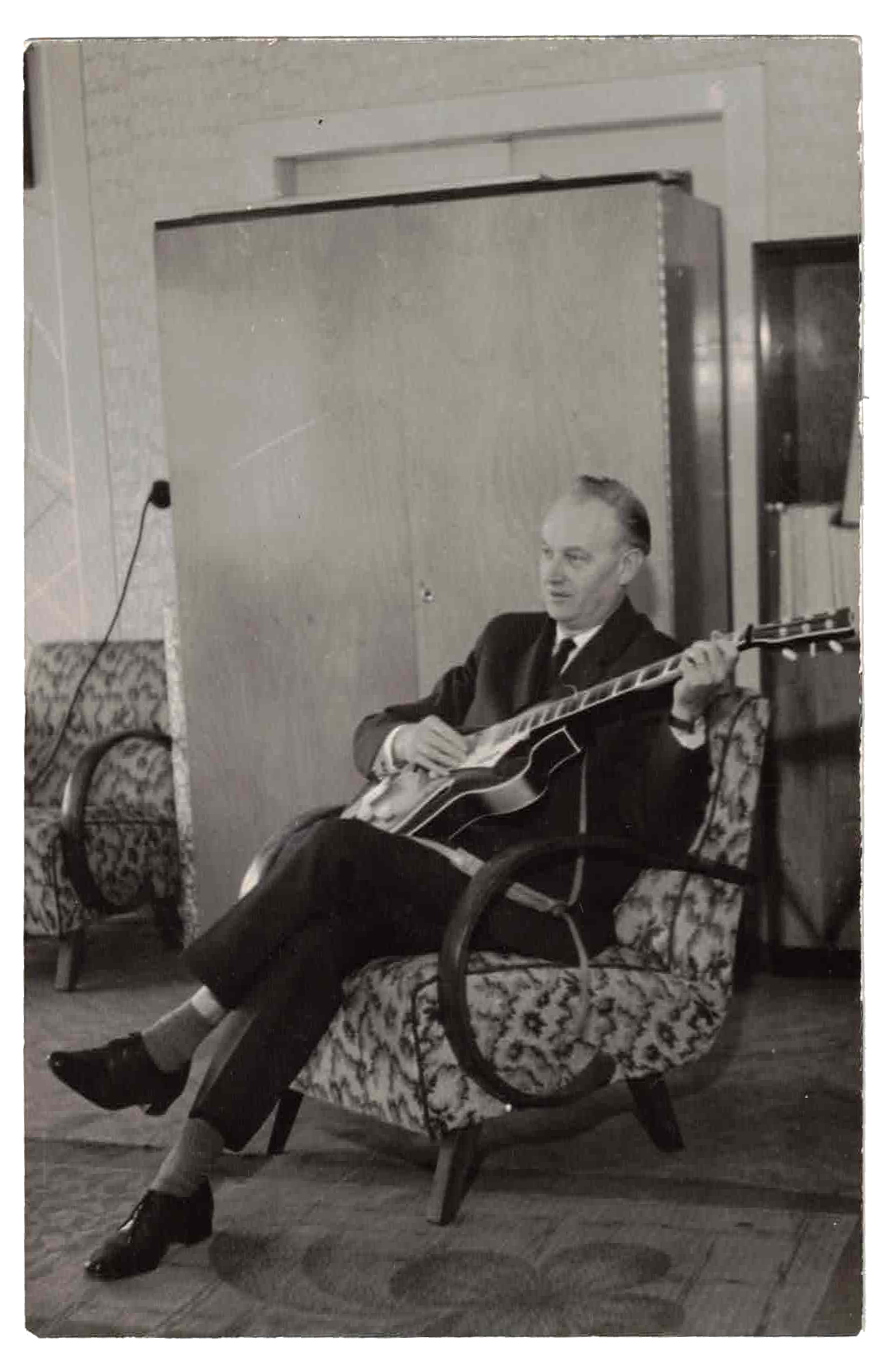 A black-and-white photo of a man sitting on a patterned upholstered armchair, playing an acoustic guitar. He is dressed in a dark suit and light shirt, with crossed legs and high heels. The room appears to be a vintage living room with another similar armchair, carpet, and a wooden cabinet in the background.