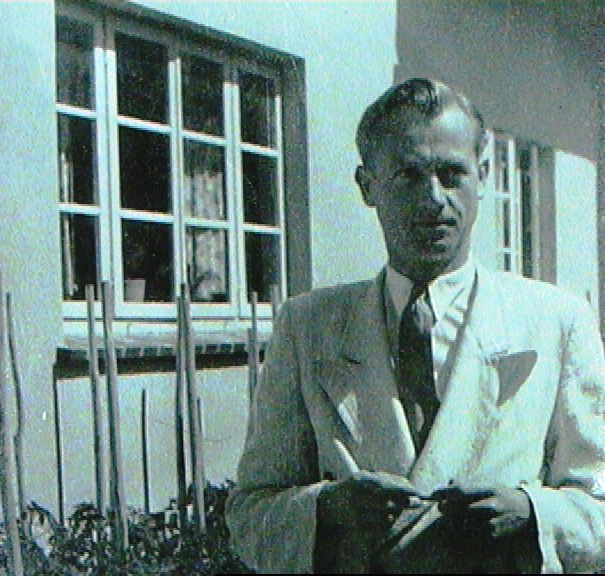 A man in a light-colored suit and dark tie, standing outside near a building with barred windows, holding a cigarette.