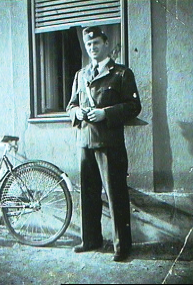 A young man standing outside near a bicycle in front of a building with a window and a partially open shutter.