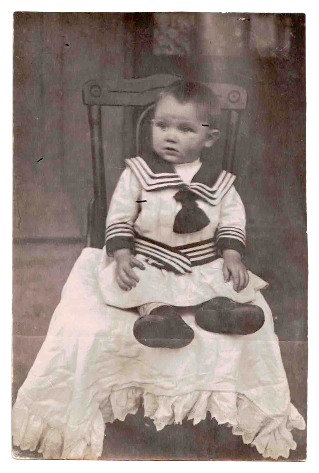 A young child sitting on a chair, dressed in a sailor-themed outfit, with a ruffled skirt and dark shoes, in a vintage black-and-white photograph.