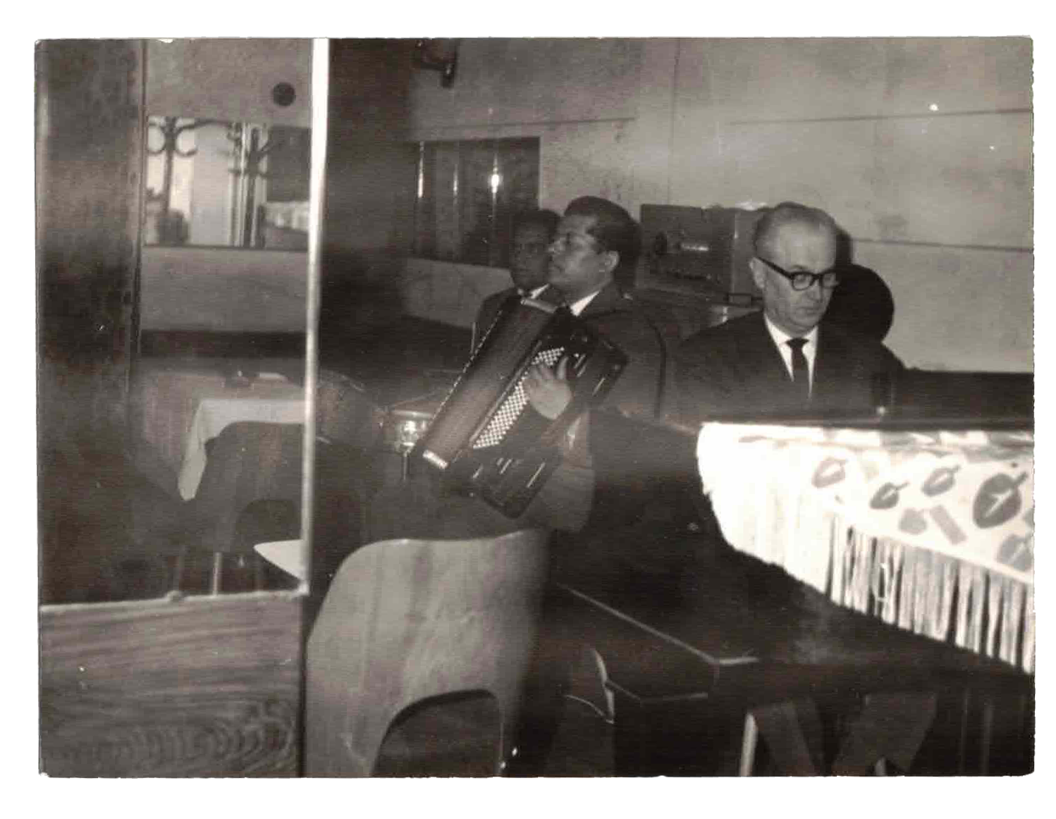 Two men in suits sitting at a table in a restaurant or cafe, with one playing an accordion and the other looking at his keyboard or piano, in a black-and-white photo.