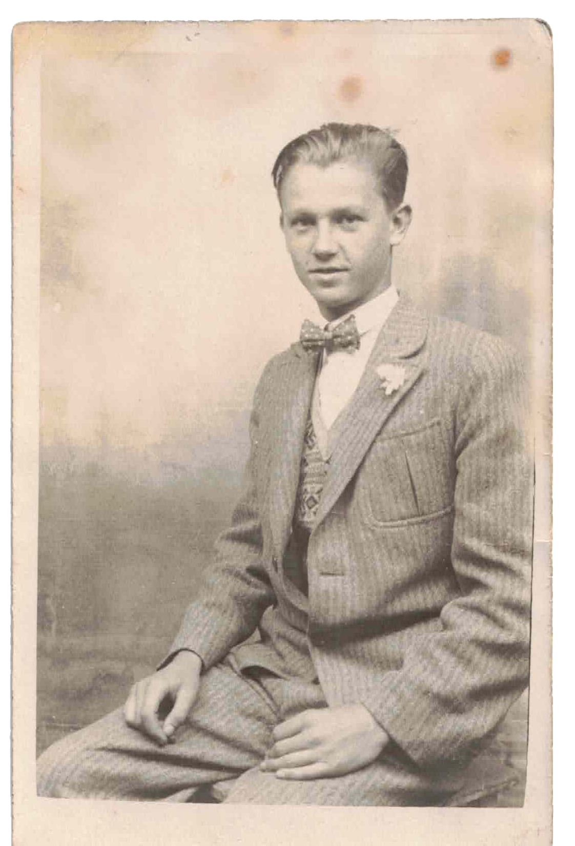 Black and white vintage photograph of a young man dressed in a suit with a bow tie, sitting with one hand resting on his lap and the other on his knee, looking at the camera.