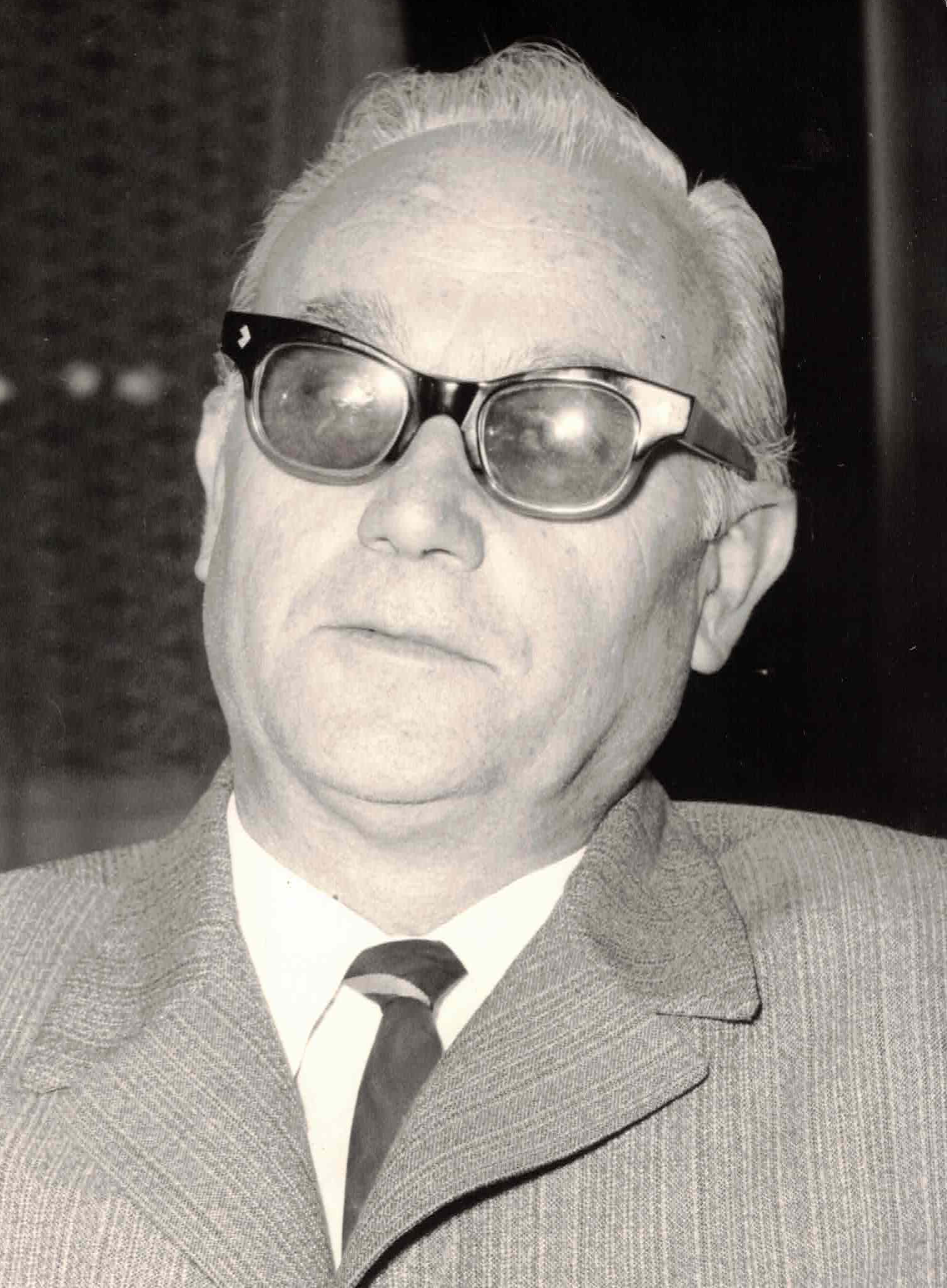 Black and white photo of an elderly man with white hair wearing glasses, a suit, dress shirt, and tie.