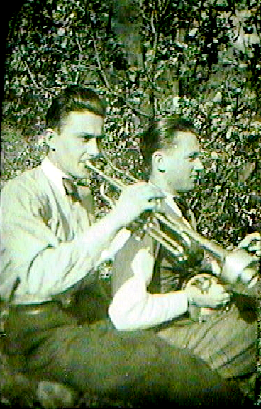 Two young men playing trumpets outdoors near bushes.