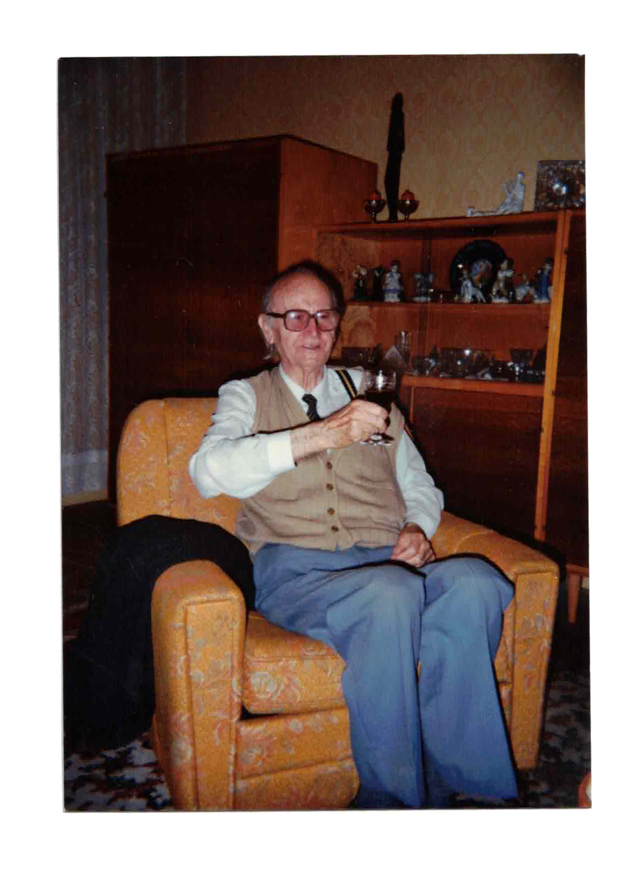 An elderly man with glasses, wearing a beige vest, white shirt, and gray pants, sitting on an orange armchair, holding a glass of dark liquid, in a room with a wooden cabinet filled with various figurines and decorative items.